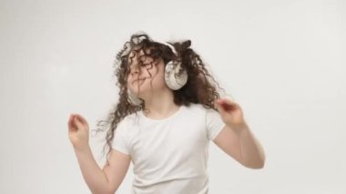 Çekici bir gülüşü olan, kablosuz kulaklıkla müzik dinleyen ve dans eden küçük, kıvırcık bir kız. Çocuğun olumlu duyguları var. İzole edilmiş, beyaz bir arka planda.