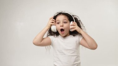 A little charming curly girl with a charming smile listens to music in wireless headphones and dances. Positive emotions of the child. Isolated, on a white background