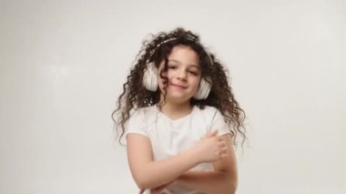 A little charming curly girl with a charming smile listens to music in wireless headphones and dances. Positive emotions of the child. Isolated, on a white background