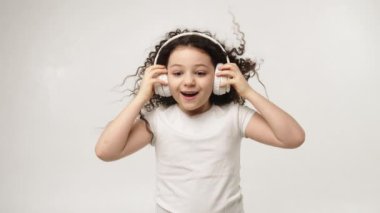 A little charming curly girl with a charming smile listens to music in wireless headphones and dances. Positive emotions of the child. Isolated, on a white background
