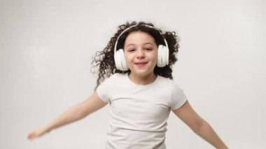 A little charming curly girl with a charming smile listens to music in wireless headphones and dances. Positive emotions of the child. Isolated, on a white background