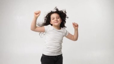 A little charming curly girl with a charming smile listens to music in wireless headphones and dances. Positive emotions of the child. Isolated, on a white background