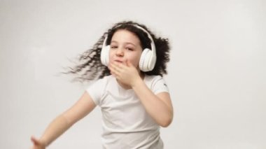 A little charming curly girl with a charming smile listens to music in wireless headphones and dances. Positive emotions of the child. Isolated, on a white background