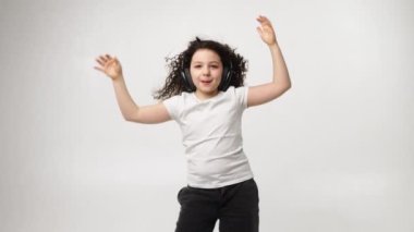 A little charming curly girl with a charming smile listens to music in wireless headphones and dances. Positive emotions of the child. Isolated, on a white background