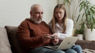 A pretty young woman shows her older mature father how to use a bank card and computer at home. An adult daughter teaches laptop apps to a focused middle-aged dad using modern technology.