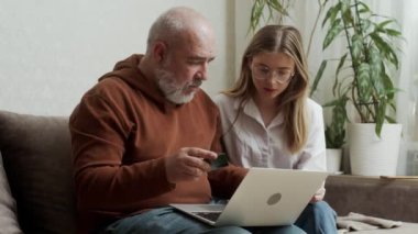 Genç ve güzel bir kadın yaşlı babasına evde nasıl banka kartı ve bilgisayar kullanılacağını gösteriyor. Yetişkin bir kız, modern teknolojiyle odaklanmış orta yaşlı bir babaya bilgisayar uygulamaları öğretiyor..