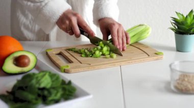 Dişi eller kereviz saplarını bıçakla küçük dilimlere ayırıyor. Mutfak masasında gri bir plastik tahta üzerinde kereviz doğrayan metal bıçak. Şef çiğ sağlıklı sebzeli vegan salatası pişiriyor..