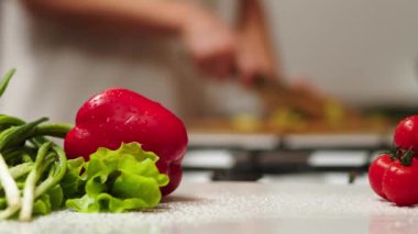the cook cuts a spring salad of tomatoes, cucumbers, greens and mixes. Woman cutting cucumber for salad on wooden cutting board on kitchen table with cucumbers, tomatoes branch and scallion around.