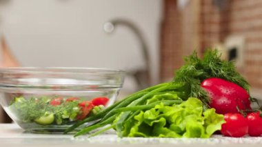 the cook cuts a spring salad of tomatoes, cucumbers, greens and mixes. Woman cutting cucumber for salad on wooden cutting board on kitchen table with cucumbers, tomatoes branch and scallion around.