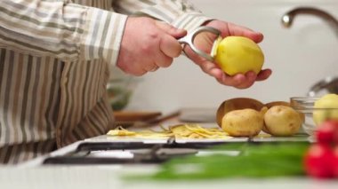 Bachelor man preparing dinner at home in the kitchen. Healthy food theme, male peels the skin on raw potatoes close-up over the table. Non-GMO vegetables from the farm. Man's hands peeling potatoes.