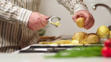 Bachelor man preparing dinner at home in the kitchen. Healthy food theme, male peels the skin on raw potatoes close-up over the table. Non-GMO vegetables from the farm. Man's hands peeling potatoes.