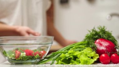 the cook cuts a spring salad of tomatoes, cucumbers, greens and mixes. Woman cutting cucumber for salad on wooden cutting board on kitchen table with cucumbers, tomatoes branch and scallion around.