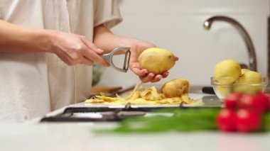 Close up view of woman in white shirt peeling a potato. Peel falling onto cutting board. Lady preparing potatoes to be cut and added to stock pot for dinner.