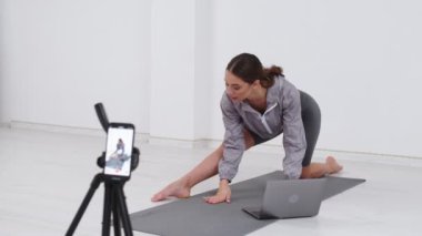 Cinematic shot of young sporty brazilian woman with athletic body wearing bra and leggings is practicing workout exercises for legs and buttocks on fitness mat with online tutorial on laptop at home.