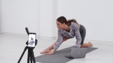 Young Girl Shooting Her Fitness Videoblog. Young Girl Conduct Remote Teaching to Training Online During Blogging. Girl Shooting a Video Blog While Doing Exercises of Stretching on Mat in Bright Room.