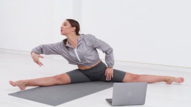 Genç atletik kadın antrenör egzersiz yapıyor, esniyor ve yoga yapıyor. Dizüstü bilgisayarla çevrimiçi fitness eğitimi.