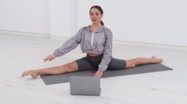 Genç atletik kadın antrenör egzersiz yapıyor, esniyor ve yoga yapıyor. Dizüstü bilgisayarla çevrimiçi fitness eğitimi.