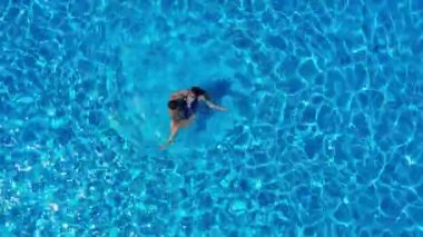 Havuzda yüzen bir çiftin uzaktan kumandalı görüntüsü. İki insan yaz tatillerinin, hafta sonlarının tadını çıkarıyor. Otelin yüzme havuzunun berrak mavi sularında dinlenen çok ırklı çiftin hava görüntüsü.