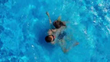 Drone view of a couple relaxing, swimming in the pool. Two people enjoy summer holidays, weekends. Aerial view of multiracial couple relaxing in clear blue water of hotel swimming pool