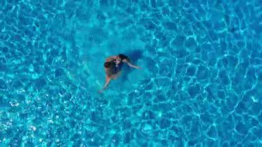Drone view of a couple relaxing, swimming in the pool. Two people enjoy summer holidays, weekends. Aerial view of multiracial couple relaxing in clear blue water of hotel swimming pool