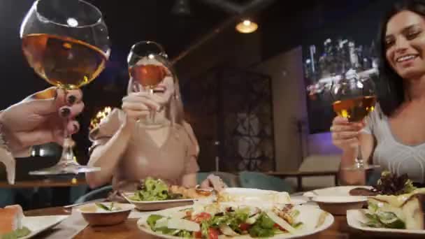 三个可爱的女朋友坐在桌旁休息 喝着酒 兴高采烈地交谈着 女孩用酒杯扣酒的玻璃杯 — 图库视频影像