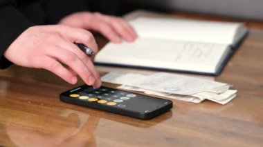 Oturma odasındaki kanepede oturan genç bir adam finansal harcamaları ve borçları hesaplamak için modern ekonomideki mali ve vergi belgelerinin gelirlerini hesaplamak için hesap makinesi kullanıyor.