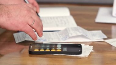 Oturma odasındaki kanepede oturan genç bir adam finansal harcamaları ve borçları hesaplamak için modern ekonomideki mali ve vergi belgelerinin gelirlerini hesaplamak için hesap makinesi kullanıyor.