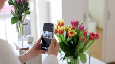Bir buket çiçeğin fotoğrafını çeken, gülümseyen genç bir kadının yan görüntüsü. Evde tatilde kırmızı lalelerle fotoğraf çeken mutlu, beyaz bir kadın. Kadınlar Günü
