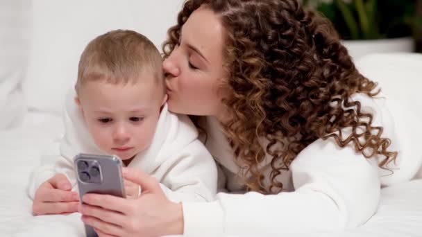 Anne Yaşındaki Küçük Oğlu Yatağa Uzanmış Cep Telefonuna Bakıyor Gülüyor — Stok video