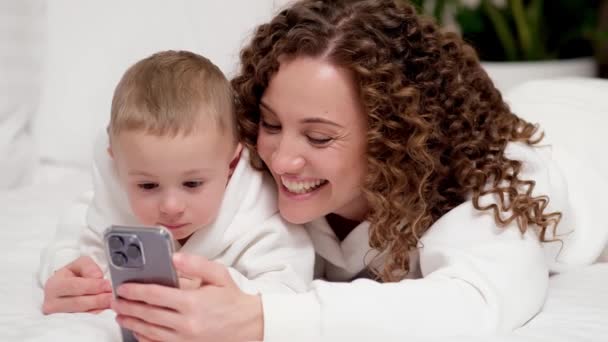 妈妈和两岁的小儿子躺在床上 看着手机 看卡通片 打视频电话 在网上玩得很开心 两代家庭使用新的应用程序 — 图库视频影像
