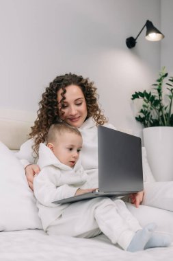 Genç Avrupalı mutlu anne ve tatlı anaokulu çocuğu dizüstü bilgisayar kullanıyor, bilgisayar ekranına bakıyor, sosyal medya sohbetinin keyfini çıkarıyor, internetten alışveriş yapıyor. Aile evde birlikte dinleniyor.