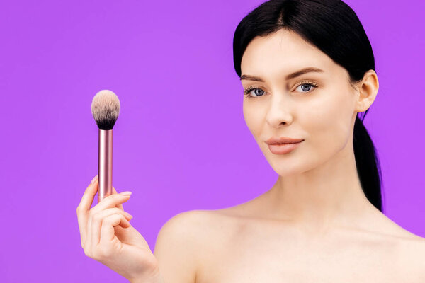 Close up portrait of attractive beautiful young topless woman standing isolated over purple background holding makeup brush