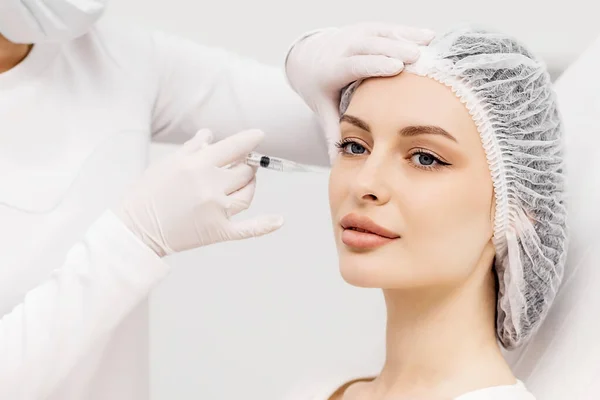 Çekici genç bir kadın canlandırıcı yüz enjeksiyonu yaptırıyor. O, klinikte sakince oturuyor. Uzman güzellik uzmanı hyaluronik asitle kadın kırışıklıklarını dolduruyor..