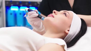 Doktor kozmetolojistin kozmetoloji ofisinde yaşlanmayı önleme prosedürü uygulaması. Tek kullanımlık şapkalı tatmin olmuş kadın kanepede uzanıyor ve dinleniyor. Aygıtla çalışmak.