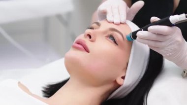 Doktor kozmetolojistin kozmetoloji ofisinde yaşlanmayı önleme prosedürü uygulaması. Tek kullanımlık şapkalı tatmin olmuş kadın kanepede uzanıyor ve dinleniyor. Aygıtla çalışmak.