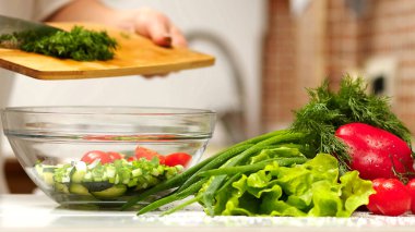 Kadın domates, salatalık, yeşillik ve karışık salata kesiyor. Salata için salatalık kesen bir kadın mutfak masasında salatalık, domates dalı ve soğanlı bir tahta kesiyor..
