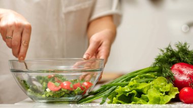 Kadın domates, salatalık, yeşillik ve karışık salata kesiyor. Salata için salatalık kesen bir kadın mutfak masasında salatalık, domates dalı ve soğanlı bir tahta kesiyor..