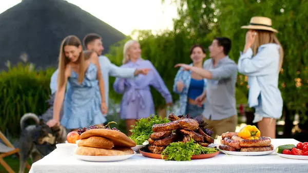 Açık hava yemek masasında leziz barbekü etleri, taze sebzeler ve salatalar. Genç bir kadın masaya koştu ve bir biftek aldı. Mutlu insanlar dans ediyor ve arka planda eğleniyorlar..