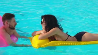 Havuzda konuşan bir çift genç aşık. Orta boy fotoğraf: bir erkek ve bir kadın şişme yüzükler ve yüzen bir şiltenin üzerinde havuzda dinlenmekten hoşlanırlar..