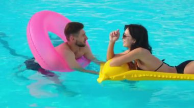 Havuzda konuşan bir çift genç aşık. Orta boy fotoğraf: bir erkek ve bir kadın şişme yüzükler ve yüzen bir şiltenin üzerinde havuzda dinlenmekten hoşlanırlar..