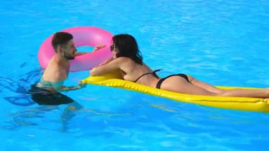 Havuzda konuşan bir çift genç aşık. Orta boy fotoğraf: bir erkek ve bir kadın şişme yüzükler ve yüzen bir şiltenin üzerinde havuzda dinlenmekten hoşlanırlar..