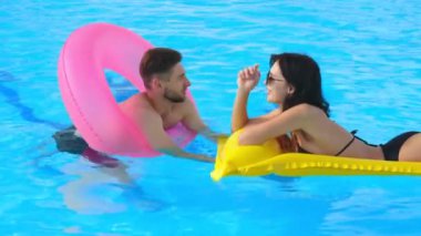 Havuzda konuşan bir çift genç aşık. Orta boy fotoğraf: bir erkek ve bir kadın şişme yüzükler ve yüzen bir şiltenin üzerinde havuzda dinlenmekten hoşlanırlar..