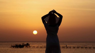 Güçlü, kendine güvenen, şapkalı güzel bir kadın. Gün batımının altında, denizde.