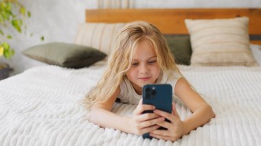 Kulaklıklar, sevimli küçük kız bir smartphone kullanarak, kameraya bakıyor ve evde kanepede yatan gülümseyen.