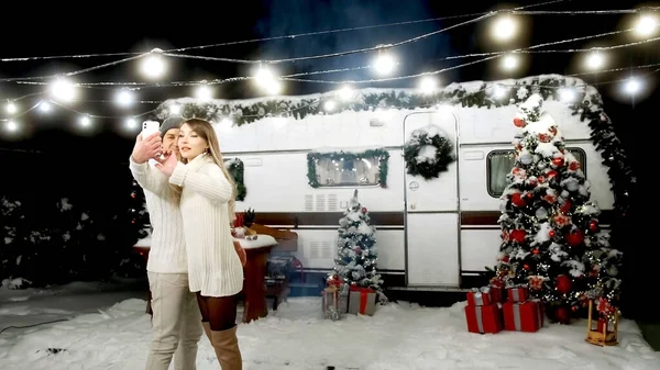 Bir adam ve bir kız, Noel ağacının yanındaki akıllı bir telefondan çelenk ve bir kampçı ile selfie çekip gülümsüyorlar. Yeni yıl ve Noel konsepti.
