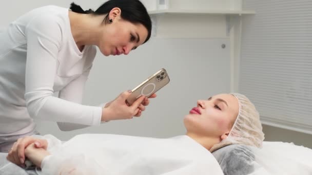 メディカルオフィス 女性医師は ヒアルロン酸 顔の矯正 化粧品の注射の後 電話で患者の写真を撮ります 高品質の4K映像 — ストック動画