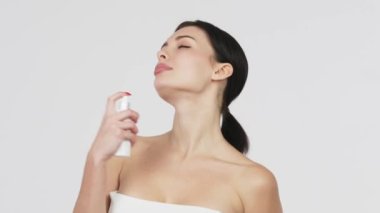 Güzel genç bir kadın banyoda dururken boynuna nemlendirici yüz sisi püskürtüyor. Güzellik, vücut bakımı ve insan konsepti. Gri arka plan üzerinden yüzüne sprey sıkan bir kadın..