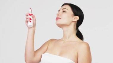 Güzel genç bir kadın banyoda dururken boynuna nemlendirici yüz sisi püskürtüyor. Güzellik, vücut bakımı ve insan konsepti. Gri arka plan üzerinden yüzüne sprey sıkan bir kadın..