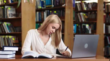 Ciddi derecede sıkı çalışan sarışın kız öğrenci webinar izliyor, çevrimiçi öğrenim kurslarını dinliyor, dizüstü bilgisayara bakıyor, üniversite kütüphanesindeki masada notlar yazıyor.