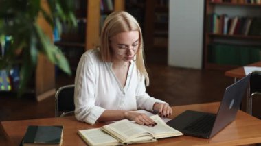 Ciddi derecede sıkı çalışan sarışın kız öğrenci webinar izliyor, çevrimiçi öğrenim kurslarını dinliyor, dizüstü bilgisayara bakıyor, üniversite kütüphanesindeki masada notlar yazıyor.